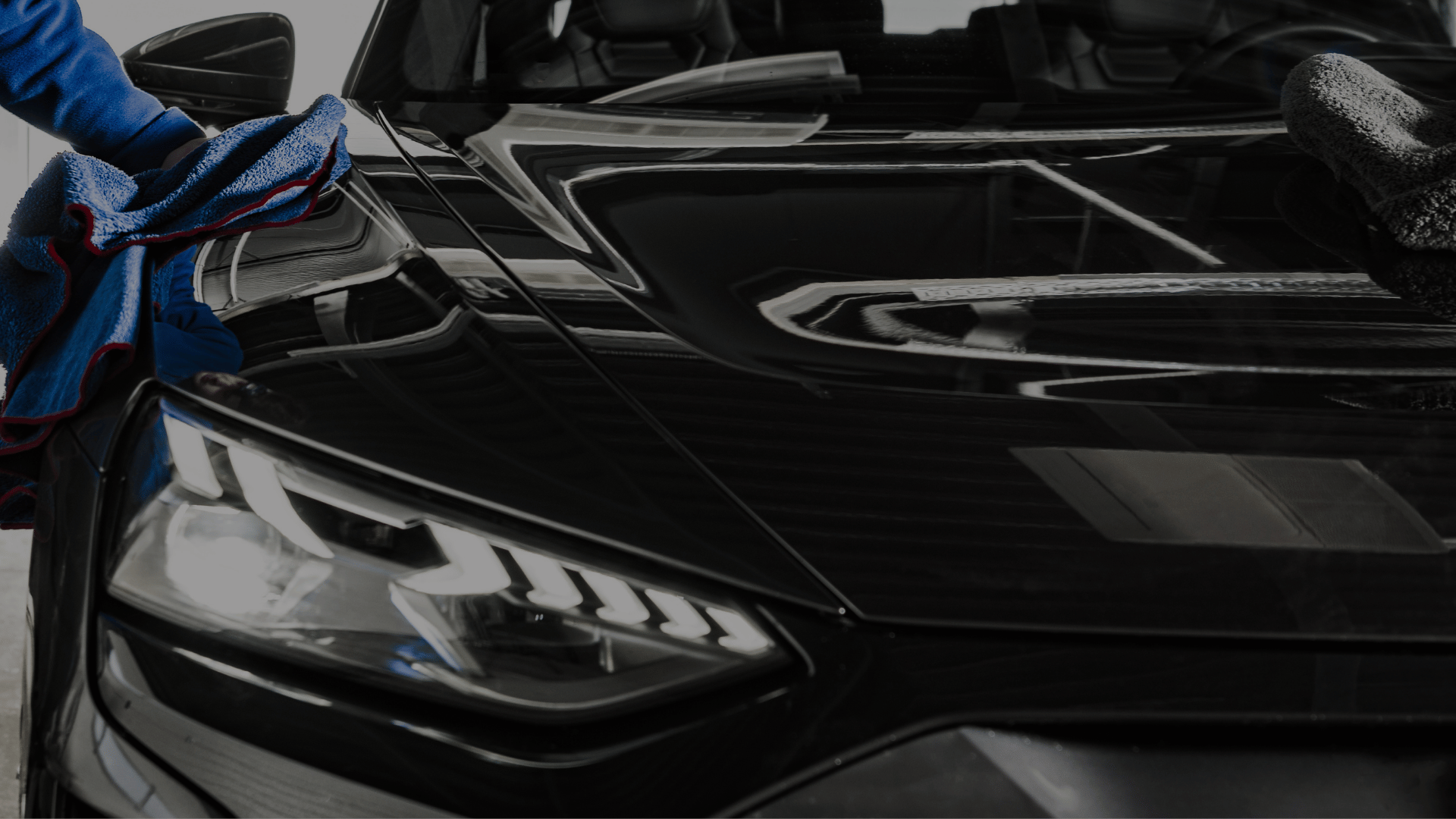 Hand wiping a black car's hood and headlight with a blue microfiber cloth in low light.