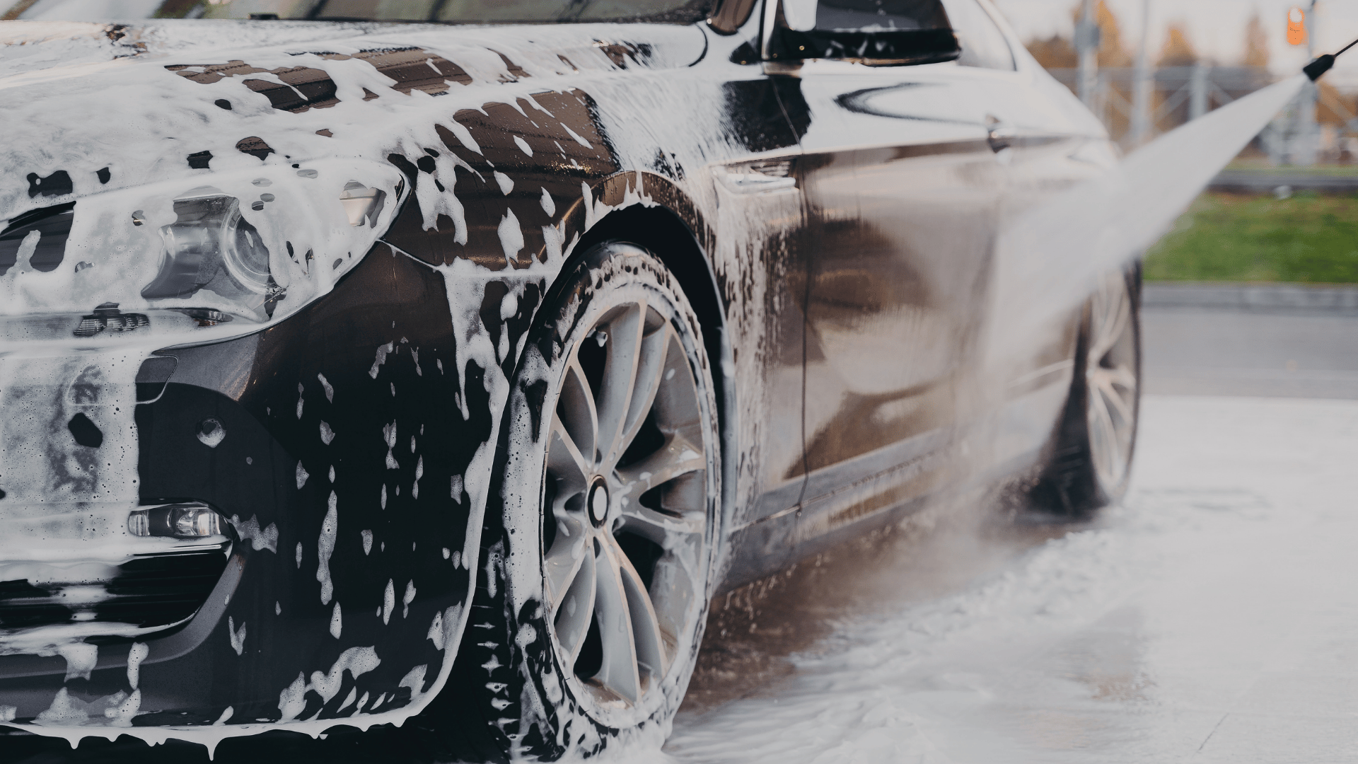 Foam-covered car being rinsed with a pressure washer, wheel and front end visible.