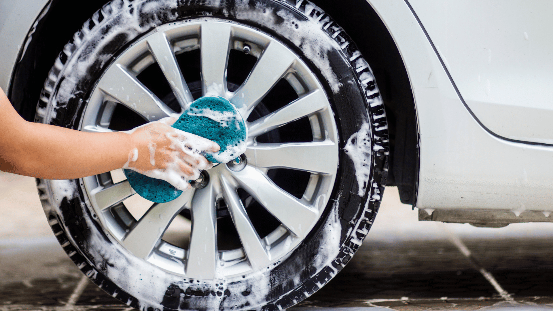 Hand scrubbing a soapy alloy wheel with a blue sponge.