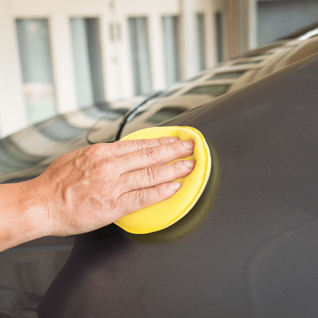 Hand applying polish to a dark car surface with a yellow foam pad.